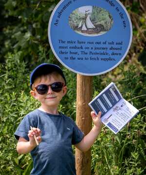 A child in sunglasses stands beside a sign, holding a pamphlet. Greenery in the background.